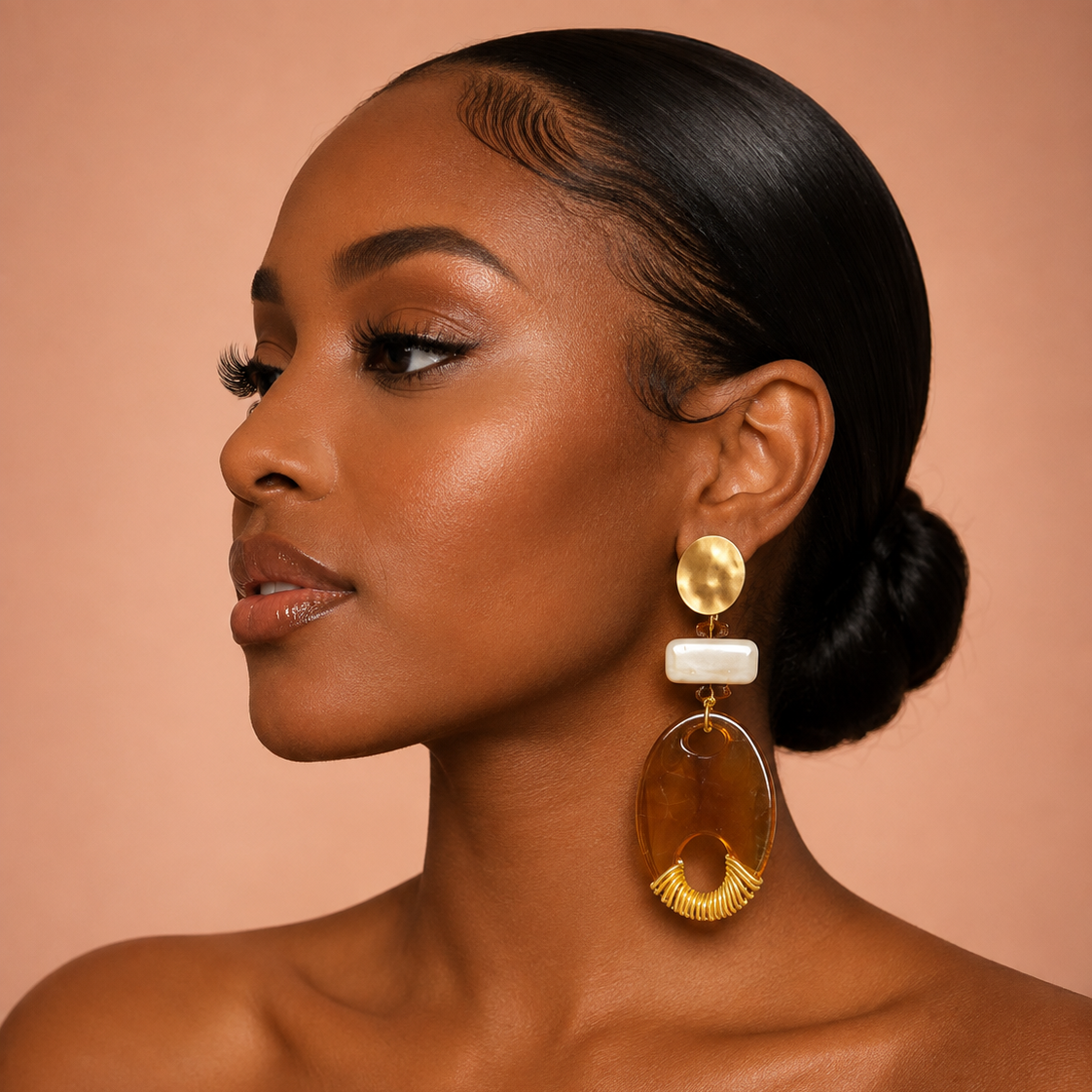 A close-up portrait of a woman wearing a large statement earring. The earring features a gold-toned circular top, a white rectangular stone, and a large teardrop-shaped amber-brown drop with a gold scalloped edge.
