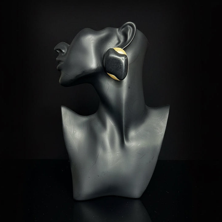 A pair of chunky black geometric drop earrings with a gold inner lining, displayed on a black mannequin bust against a gray subway tile wall.