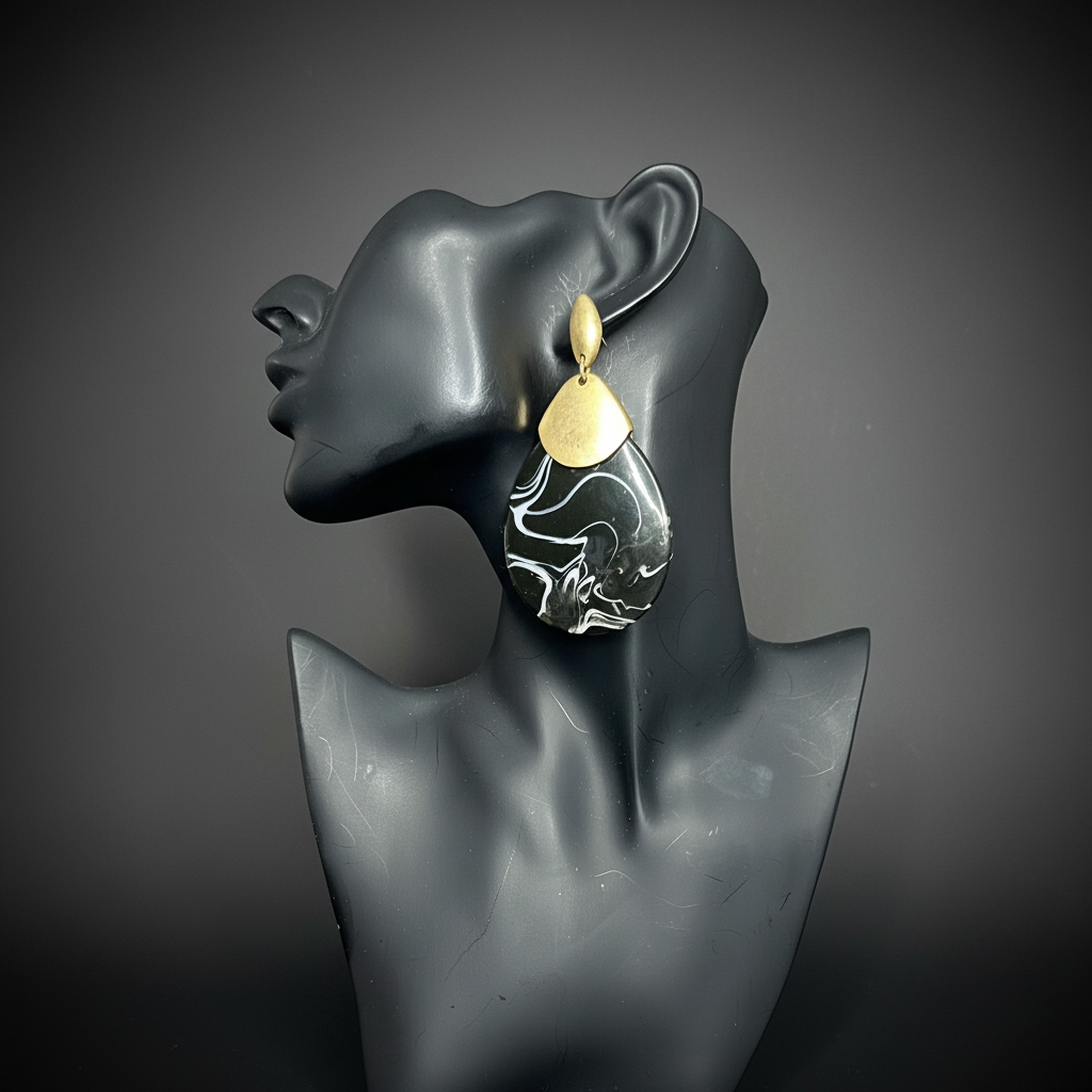 A pair of large sculptural drop earrings displayed on a matte black mannequin bust against a dark gray gradient background. earring is matte gold with a teardrop-shaped dangle, abstract marbled swirl pattern.