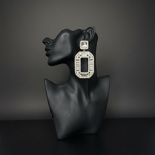 A large rectangular statement earring with a silver-toned frame and clear rhinestones, displayed on a black mannequin bust against a gray subway tile wall.