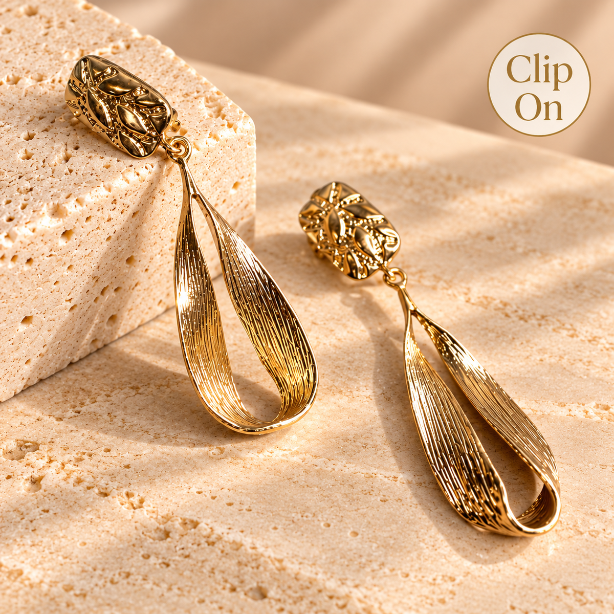 A pair of gold drop earrings with textured, leaf-like teardrop pendants and floral stud tops, displayed on a beige stone surface with a 'Clip On' badge in the top right corner.