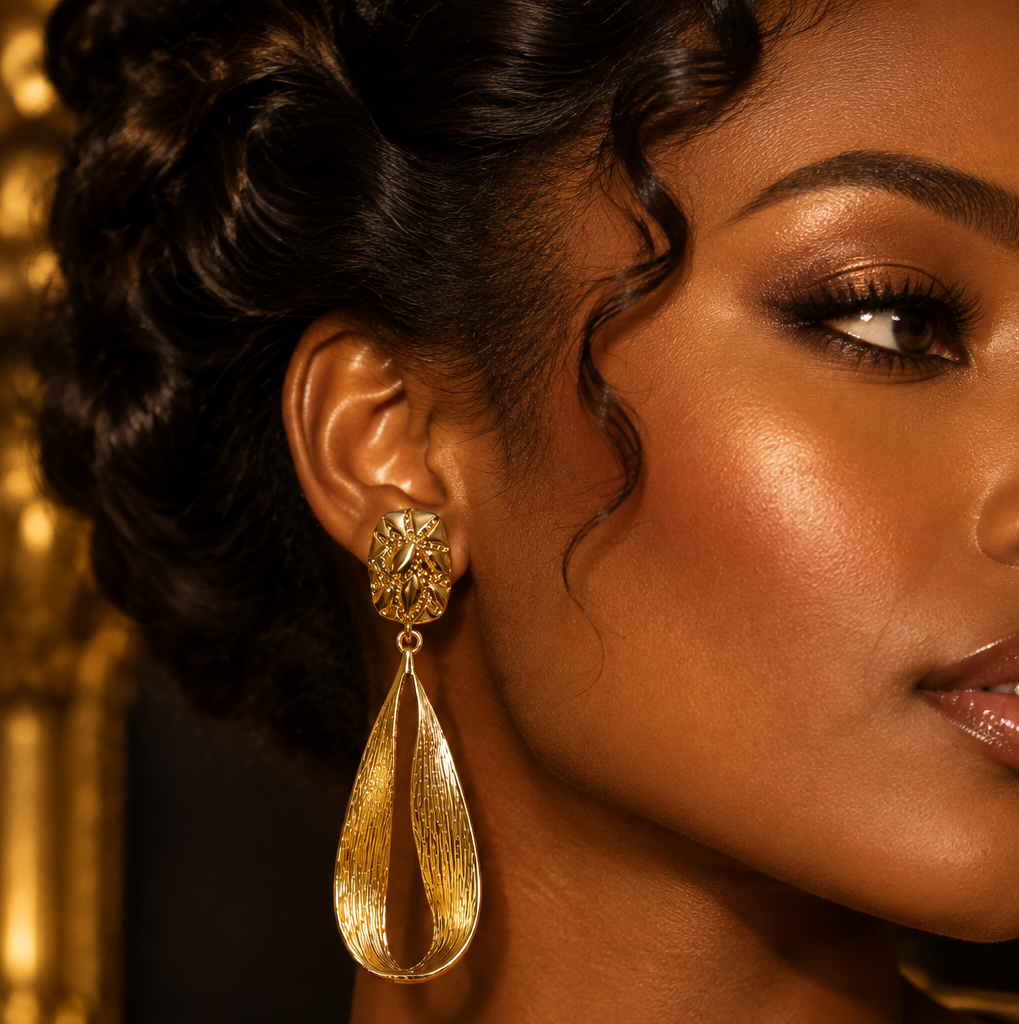 A close-up of a woman's ear wearing a large textured gold dangle earring with a teardrop-shaped drop and a floral stud, set against a warm, softly blurred background.