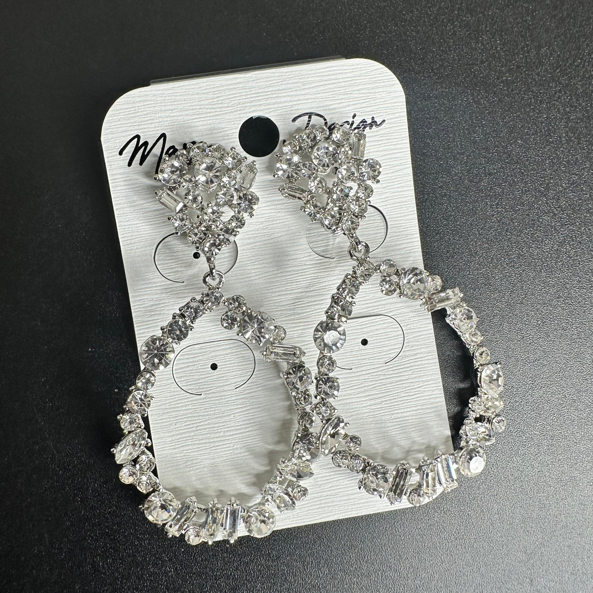 A pair of silver-tone hoop earrings with clear rhinestone embellishments, displayed on a white branded card against a dark gray background.