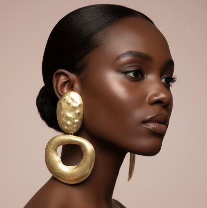 A woman wearing A pair of chunky gold-tone statement earrings with one oval textured drop and one larger open circular drop, displayed  against a nude wall