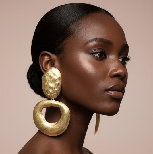A woman wearing A pair of chunky gold-tone statement earrings with one oval textured drop and one larger open circular drop, displayed  against a nude wall