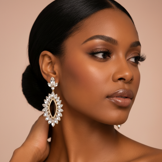 A close-up portrait of a woman wearing a large, ornate teardrop-shaped dangle earring with clear crystals and gold-toned metal details. The earring is displayed on her ear, with her other hand resting near her neck against a solid, warm beige background.