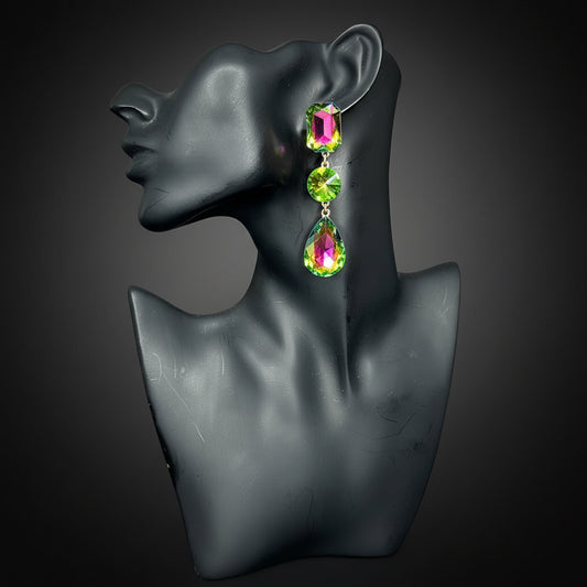 A pair of long drop earrings with iridescent green and pink teardrop-shaped crystals in gold-toned settings, displayed on a black female bust against a dark background 