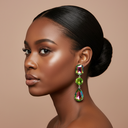 A woman in profile wearing a pair of dramatic drop earrings with green and iridescent marquise-cut crystals, set against a solid warm beige background.