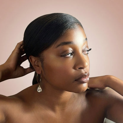 Woman wearing Gold earring with a clear teardrop-shaped gemstone  against a pink background