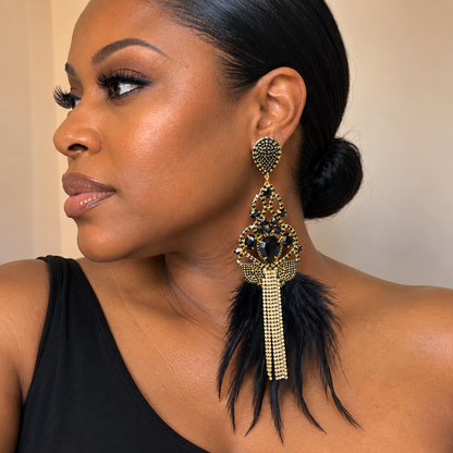 A woman wearing a large statement dangle earring with gold-tone metal, dark blue stones, and black feathers, shown in a close-up portrait against a plain beige wall.