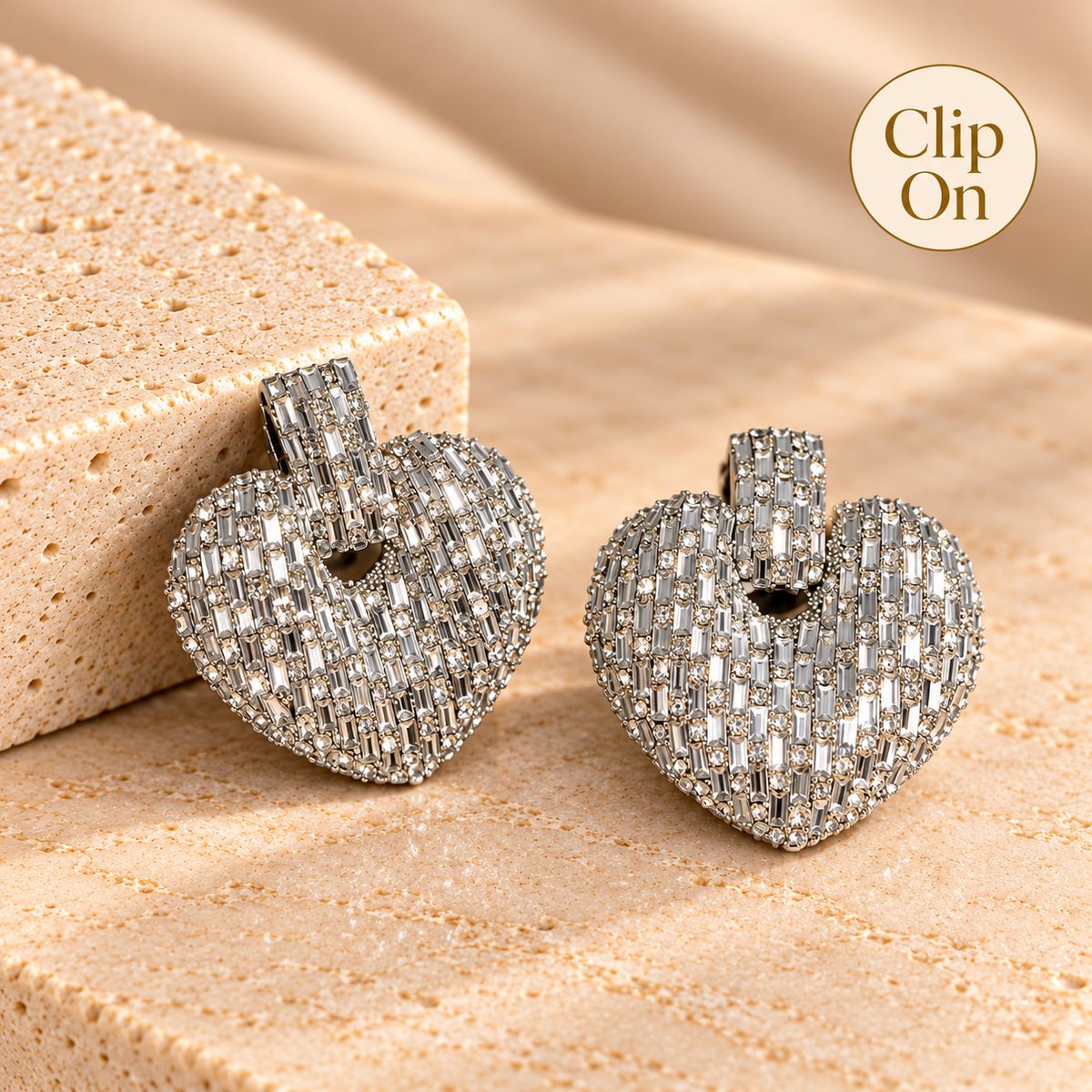 A pair of heart-shaped clip-on earrings covered in sparkling clear stones, displayed on a textured beige surface next to a sponge block. A circular 'Clip On' badge is in the top right corner.