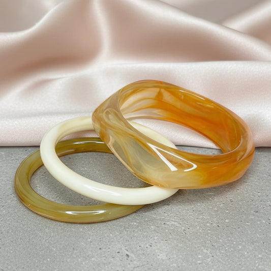 Three translucent bracelets in orange, green and white on a dark surface with a tiled wall background.