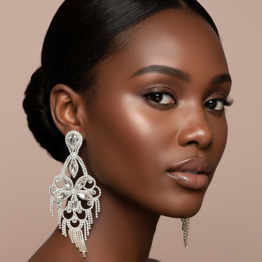Woman wearing A long silver-toned statement dangle earring with intricate filigree and rhinestone-like details, displayed against a blush background 