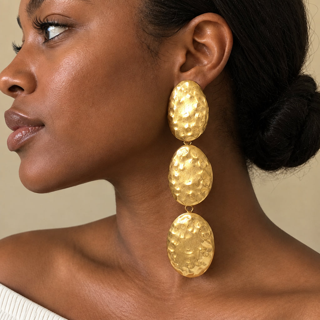 A close-up side profile of a woman wearing a long, three-drop statement earring made of textured gold-toned metal discs.
