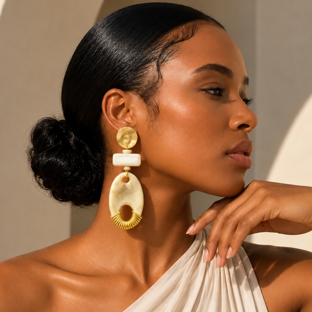 A woman in a draped cream dress wearing large statement earrings with gold and cream-colored organic shapes, photographed in warm sunlight against a beige wall.
