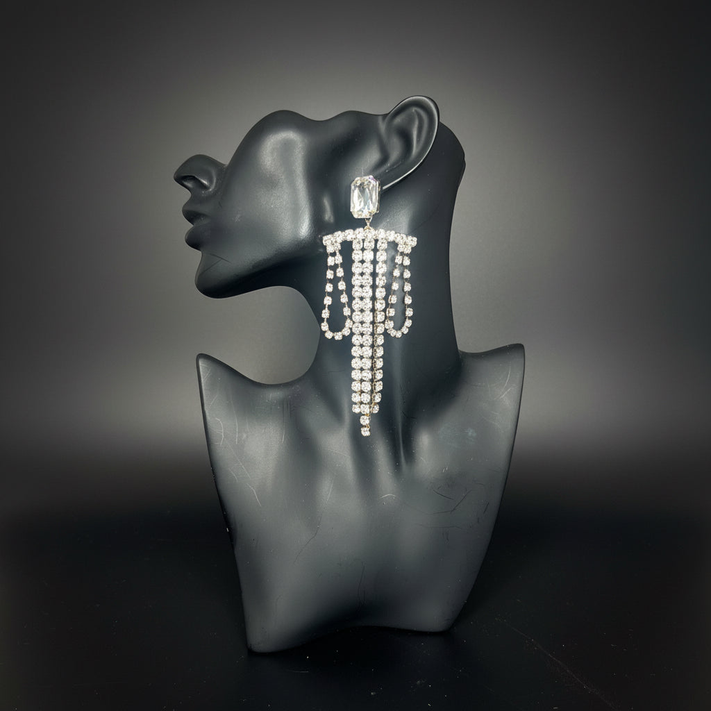 A long, dangling statement earring covered in clear rhinestones on a black mannequin bust, set against a dark background 