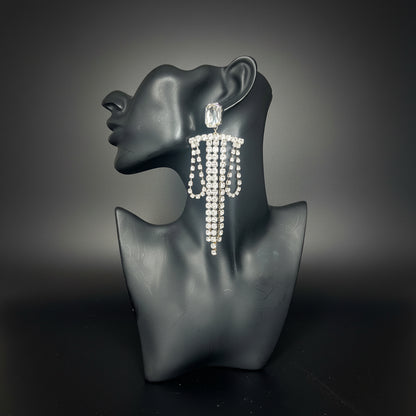 A long, dangling statement earring covered in clear rhinestones on a black mannequin bust, set against a dark background 