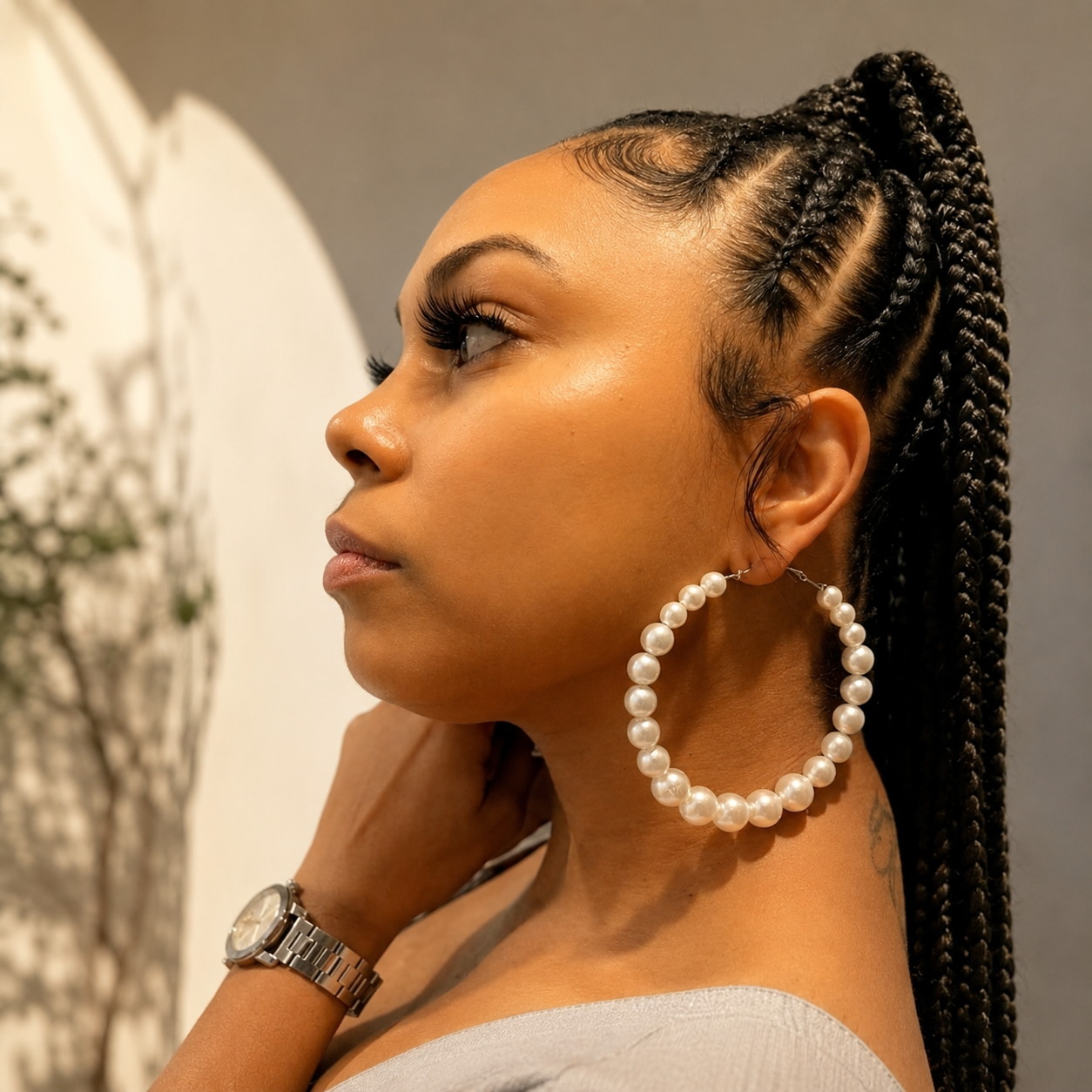 A close-up side profile of a woman wearing large, chunky hoop earrings made of white freshwater pearls. She has dark cornrow braids in her hair and is wearing a silver watch on her wrist, set against a soft-focus indoor background.