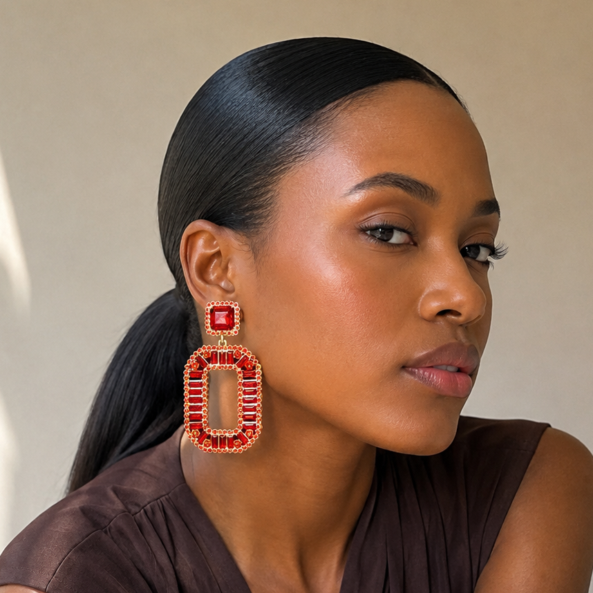 A woman with her hair in a ponytail wearing a brown top and large red beaded geometric drop earrings, posed against a neutral beige wall with soft window shadows.