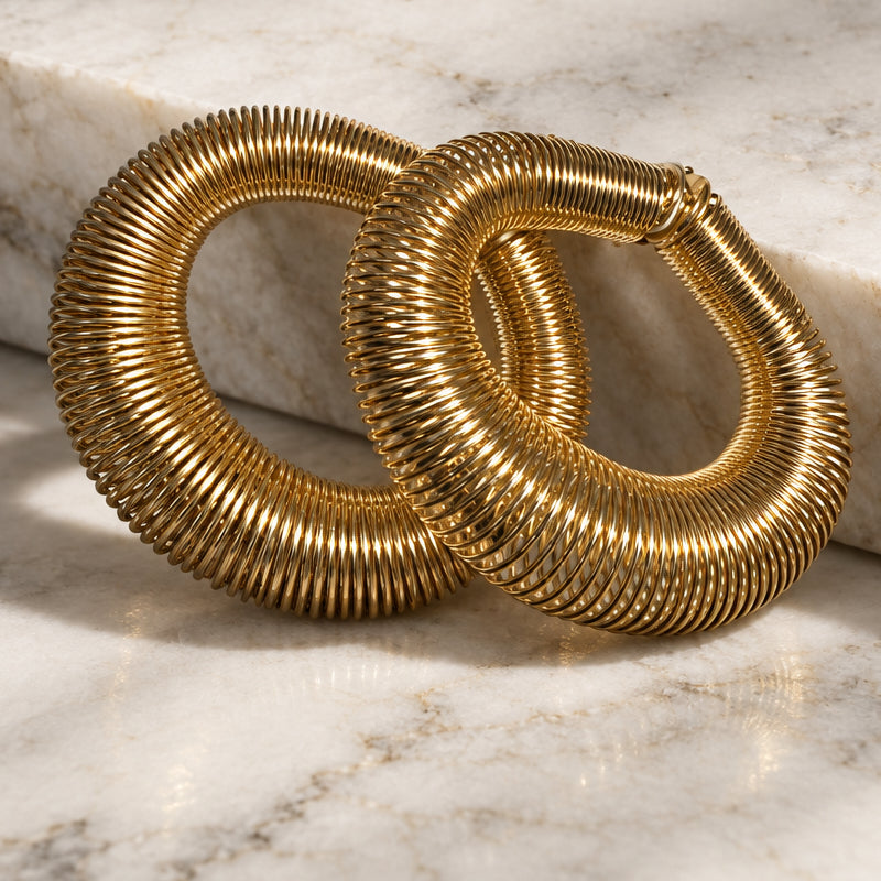 Two oversized ribbed gold hoop earrings displayed on a neutral cream marble surface.