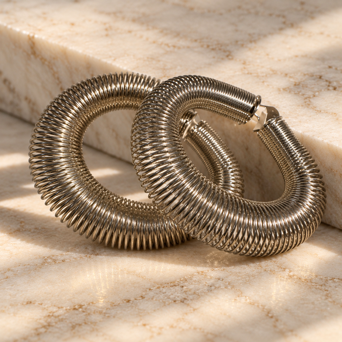 A pair of large, textured silver hoop earrings with a coiled, snake-like ribbed design, resting on a beige marble surface with soft shadows.