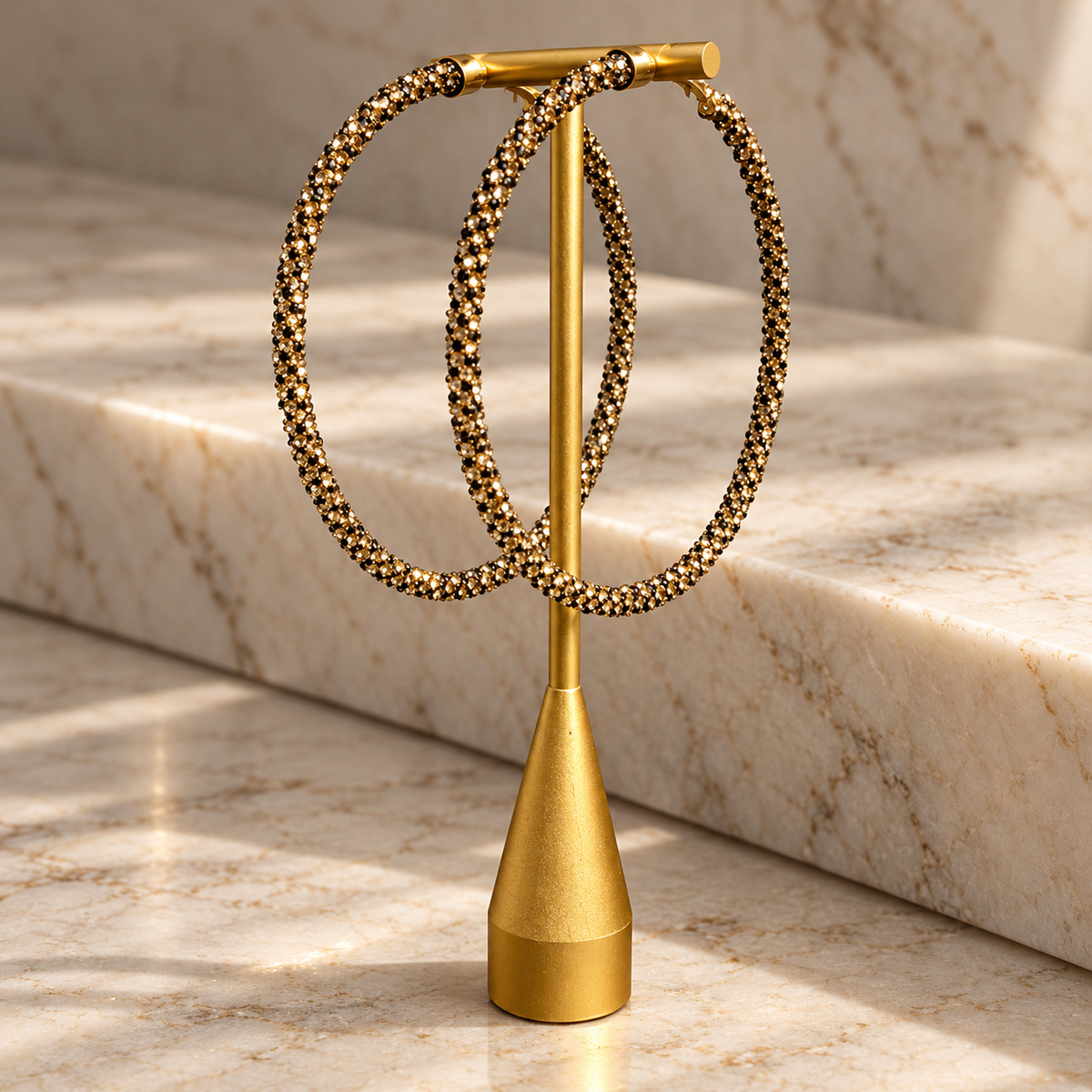 A pair of large gold hoop earrings with textured beaded edges, displayed on a gold stand against a marble background.