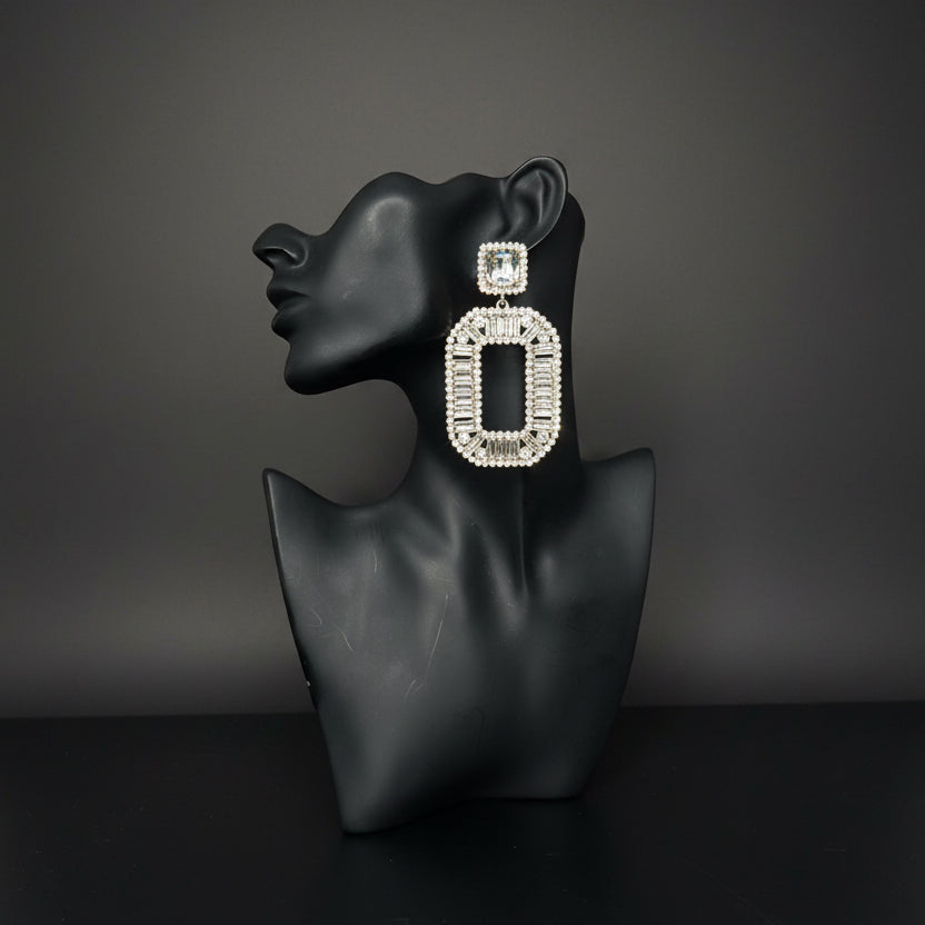 A large rectangular statement earring with a silver-toned frame and clear rhinestones, displayed on a black mannequin bust against a gray subway tile wall.