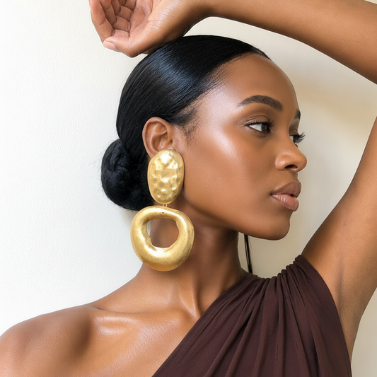A woman with her hair in a low bun models a pair of large, chunky gold statement earrings with textured circular and oval shapes. She is wearing a brown draped one-shoulder top against a plain, warm off-white background.