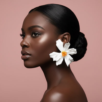 A woman in profile with dark hair in a bun wearing a large white flower earring with a yellow-orange center, set against a solid dusty pink background.