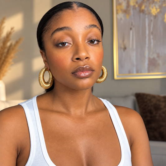A woman with slicked-back hair wearing large textured gold hoop earrings and a white ribbed tank top, photographed in a softly lit indoor setting with framed wall art and home decor in the background.