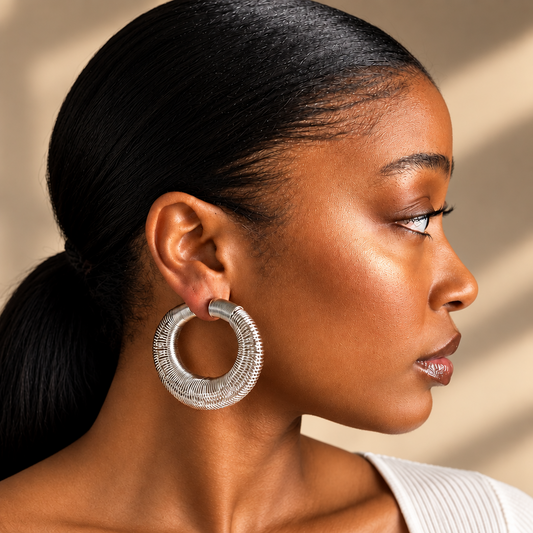 A close-up side profile of a woman with dark hair in a ponytail, wearing a large textured silver hoop earring. The background is a soft beige wall with warm light shadows.