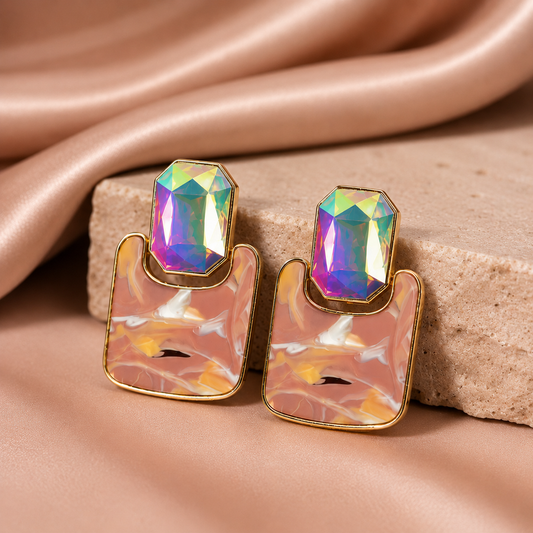 A pair of gold-tone statement earrings with iridescent, rainbow-flecked rectangular gemstones on top of blush pink marbled resin, displayed on a beige stone block with draped satin fabric.